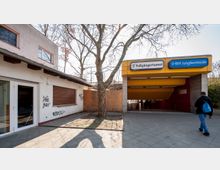 Eingang zu einem Fußgängertunnel und der U-Bahn-Station Jungfernheide, gekennzeichnet durch ein gelbes, überdachtes Vordach mit Schildern. Links ist ein Gebäude mit geschlossenen Rollläden und Graffiti zu sehen, im Vordergrund steht ein kahler Baum.