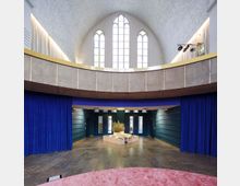 Innenraum einer modern umgestalteten Kirche mit gewölbter Decke, großen gotischen Fenstern und einer umlaufenden Galerie mit Betonelementen. Der Boden ist aus dunklem Stein, blaue Vorhänge und eine zentrale Pflanzendekoration betonen die moderne Gestaltung.