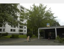 Wohngebäude und U-Bahn-Eingang in einer urbanen Umgebung. Links steht ein mehrstöckiges Wohnhaus mit weißen Fassadenplatten und Balkonen, im Vordergrund ein grüner Rasen und Bäume; rechts ein Eingang zur U-Bahn-Station "Hansaplatz" mit einem blauen Schild und der Aufschrift "U9".