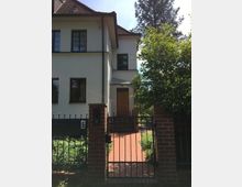 Ein zweistöckiges Wohnhaus mit weißer Fassade und dunkelbraunem Ziegeldach, umgeben von einem Garten. Im Vordergrund führt ein Weg aus roten Ziegelsteinen durch ein schmiedeeisernes Tor mit Backsteinpfeilern zur hölzernen Eingangstür.