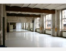 Ein Tanzstudio mit hohen Decken, Holzbalken und unverputzten Ziegelwänden. Der Raum hat große Fenster, Spiegel entlang einer Wand und Holzstangen, die als Ballettstangen dienen, sowie einen glänzenden weißen Boden.