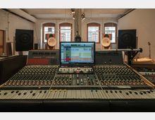 Tonstudio mit einem großen Mischpult im Vordergrund, auf dem ein Monitor mit Audiosoftware steht. Im Hintergrund sind hohe Fenster, Lautsprecher und Hörner für die Audiowiedergabe zu sehen.