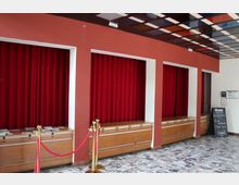 Ein Eingangsbereich mit roten Vorhängen in drei Nischen, hölzernen Tresen darunter und goldenen Absperrpfosten mit roten Kordeln. Der Fußboden ist mit gemusterten Fliesen gestaltet, und an der Seite steht eine Tafel mit Begrüßungstext.