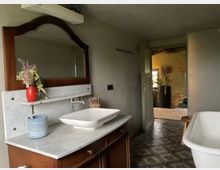 Badezimmer mit einem Waschtisch aus Holz und einer weißen Marmorplatte, darauf ein rechteckiges Keramikwaschbecken mit Armatur. Im Hintergrund ein offener Durchgang zu einem Raum mit Pflanzen und einer gelben Wand.