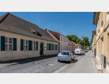 Straßenansicht mit historischem Gebäude links mit weißen Fassaden, blauen Fensterläden und Ziegeldach. Rechts geparkte Autos entlang einer gepflasterten Straße, flankiert von Gehwegen und Laternen; im Hintergrund Bäume unter blauem Himmel.