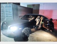 Ein schwarzes Luxusauto mit eingeschalteten Frontscheinwerfern steht auf einem Betonboden, eine Tür auf der Beifahrerseite ist geöffnet. Im Hintergrund sind Container und eine Betonwand sichtbar, teils in gedämpftem Licht und mit einem roten Lichteffekt.