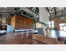 Großer Veranstaltungsraum mit Holzboden und hohen Backsteinwänden. Sichtbar sind eine Bühne mit einem Rednerpult, eine große Leinwand, Stehtische mit roten Tischläufern und ein modern gestalteter Würfel aus rostfarbenem Metall an der Stirnseite. Im Hintergrund sind hohe Fenster mit Rundbögen und eine breite, gläserne Ausgangstür.