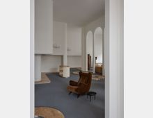 Innenraum mit modernem Design, geprägt von weißen Wänden, hohen Rundsäulen und bogenförmigen Durchgängen. Im Vordergrund stehen ein brauner, gepolsterter Sessel und ein kleiner Beistelltisch auf einem grauen Teppichboden; ergänzend sind helle Holzelemente und minimalistische Möbel sichtbar.