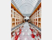 Großer, modern gestalteter Innenhof mit einem Glasdach in gewölbter Gitterstruktur, flankiert von symmetrischen gelben Gebäudefassaden mit rechteckigen Fenstern. In der Mitte befindet sich eine geschwungene, silbern glänzende Skulptur, die auf einer spiegelnden Glasfläche zu stehen scheint.