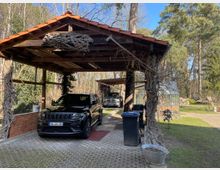 Carport mit Ziegeldach, unter dem zwei Autos parken, umgeben von Bäumen und Grünfläche. Im Hintergrund ist ein Gewächshaus und eine ländliche Gartenumgebung zu sehen.