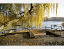Steinufer mit einer rechteckigen Plattform und Geländern, umgeben von herabhängenden Zweigen einer Trauerweide. Im Hintergrund ist ein ruhiger See mit Bäumen am Ufer zu sehen.