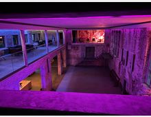 Großer, offener Innenraum mit unverputzten Backsteinwänden, einer Galerie mit Glasgeländern und leuchtender rosa und violetter Beleuchtung. Im oberen Bereich sind Sitzmöbel und ein kunstvoll bemaltes Wandbild sichtbar.