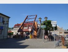 Eine kleine Klappbrücke aus Metall mit seitlichen Treppen, umgeben von gepflasterten Wegen und Wegweisern. Im Hintergrund sind Gebäude mit roten Dächern und eine Baumreihe sichtbar, klare Wetterbedingungen prägen die Szene.