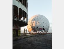 Eine verlassene Radaranlage mit einer großen, weißen, geodätischen Kuppel, die Graffiti trägt, und einem benachbarten baufälligen Gebäude mit einer beschädigten Fassade und ebenfalls Graffiti. Der Boden ist rissig und von Betonplatten bedeckt, umgeben von einem weiten, offenen Himmel.