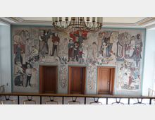 Großer Wandmosaik im Innenraum eines Gebäudes, das verschiedene Szenen des alltäglichen Lebens darstellt, mit Figuren, die Aktivitäten wie Arbeit, Familie und Gemeinschaftsszenen zeigen. Im unteren Bereich befinden sich drei hölzerne Türen, darüber hängt ein großer Kronleuchter.