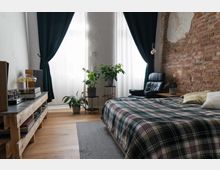 Schlafzimmer mit gemauertem Ziegelwandteil, einem Bett mit karierter Tagesdecke, einem schwarzen Ledersessel und mehreren Zimmerpflanzen auf Pflanzenständern. Große Fenster mit durchsichtigen weißen Vorhängen und dunklen, schweren Gardinen lassen Tageslicht herein. Ein Holzregal mit Schallplatten und einer Lampe steht an der Wand.