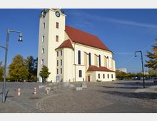 Gelbe Kirche mit rotem Satteldach und hohen Rundbogenfenstern, daneben ein Kirchturm mit Turmuhr, umgeben von einem gepflasterten Platz mit vereinzelten Laternen und Bäumen im Hintergrund.