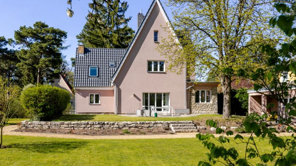 Einfamilienhaus mit steilem Giebel und dunklem Ziegeldach, davor eine kleine Terrasse und eine niedrige Steinmauer mit Stufen. Das Haus steht in einem großen Garten mit Rasenfläche, umgeben von hohen Bäumen und Sträuchern.