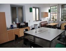 Ein einfacher Gemeinschaftsraum mit weißen Wänden, mehreren Tischen, schwarzen Kunststoffstühlen und einer Holzeckbank. Im Hintergrund befinden sich Fenster mit Blick nach draußen, ein Schrank, ein kleiner Elektroherd und diverse Pflanzen auf der Fensterbank.