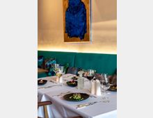 Elegantes Restaurant mit gedecktem Tisch, weißer Tischdecke, schwarzen Tellern mit dekorativen Speisen, Weingläsern und Karaffe; im Hintergrund eine grüne Polsterbank und ein abstraktes, blau-goldenes Wandgemälde.