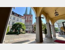 Blick durch Arkaden auf einen gepflasterten Platz mit Bäumen, modernen Gebäuden und der historischen Nikolaikirche mit ihren zwei markanten Türmen im Hintergrund. Die Szene ist von Tageslicht beleuchtet.