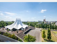 Das Bild zeigt das Tempodrom in Berlin, ein weißes, zeltförmiges Gebäude mit spitzen Dachlinien, umgeben von einer weitläufigen Holzterrasse und grünen Bäumen. Im Hintergrund sind ein Park, einige Sportplätze sowie die Stadtlandschaft mit hohen Gebäuden sichtbar.