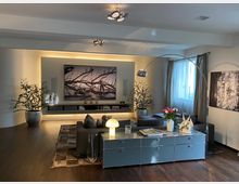 Modernes Wohnzimmer mit Holzboden, einem grauen Sofa, großen Pflanzen in Töpfen und dekorativen Kunstwerken an den Wänden. Ein Sideboard mit Lampe, Blumen und Dekorationsobjekten steht im Vordergrund, während ein großes, beleuchtetes Wandbild den Raum akzentuiert.