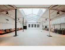 Heller, offener Raum in einem industriellen Stil, mit weißen Wänden und Säulen, einem Dach aus Glasplatten sowie Reihen von Holzbänken entlang der Seitenwände. Der Boden ist aus hellem Beton, und an den Wänden befinden sich Fenster mit Metallgittern.