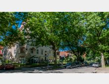 Mehrstöckiges Wohngebäude im klassischen Baustil mit großen Fenstern und verzierten Fassadenelementen, umgeben von Bäumen. Im Vordergrund führt eine mit Autos gesäumte Kopfsteinpflasterstraße entlang, im Hintergrund sind weitere Häuser und Gärten zu sehen.