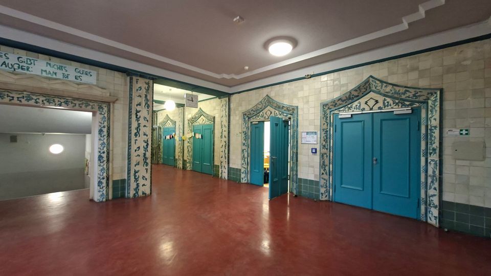 Großer Innenflur mit rötlichem Boden, hell gefliesten Wänden und mehreren türkisfarbenen Türen, die von dekorativen, gemusterten Fliesenrahmen eingefasst sind. An der Decke hängen runde Leuchten, links führt ein offener Durchgang in ein Treppenhaus.
