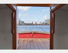 Blick von einem Boot durch eine Türöffnung auf die Spree und die Oberbaumbrücke in Berlin, umgeben von modernen und historischen Gebäuden. Im Hintergrund sind Baukräne und der Berliner Fernsehturm zu sehen.