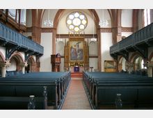 Innenraum einer historischen Kirche mit Holzbankreihen auf beiden Seiten eines Mittelgangs. Im Vordergrund sind blau gestrichene Holzbänke zu sehen, dahinter ein kunstvoller Altar mit einem Gemälde und einer bunten Rosettenfenster darüber, flankiert von roten Backsteinbögen und Emporen.
