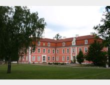 Ein zweistöckiges Herrenhaus in rosa Putzfassade mit weißen Fensterrahmen und einem roten Ziegeldach, umgeben von einer Rasenfläche und vereinzelt stehenden Bäumen. Das Gebäude hat symmetrische Fensteranordnungen, einen zentralen Giebel mit Uhr und einen Hauptzugang mit Veranda.