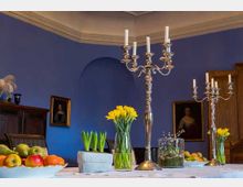 Ein Speisesaal mit blau gestrichenen Wänden, eingerahmten Porträts und einer weißen Tischdecke. Der Tisch ist dekoriert mit Kerzenhaltern, gelben Narzissen, Obstschalen und anderen Pflanzen.