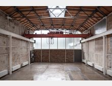 Innenansicht einer geräumigen, leerstehenden Industriehalle mit einer Konstruktion aus Mauerwerk und Sichtmetallträgern. Eine rote Laufkatze hängt an einem Stahlträger, und die Halle hat Oberlichter und Fenster mit Milchglas, die diffuses Licht hereinlassen.