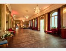 Eleganter Saal mit hohen Decken, verzierten Kronleuchtern, großen Bogenfenstern mit roten Vorhängen und einer offenen Doppeltür. Der Raum ist mit antiken Möbeln, roten Polstersofas und Blumenarrangements dekoriert, die Atmosphäre wirkt klassisch und einladend.