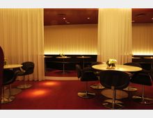 Innenraum einer Lounge mit modernem Design: Rundtische mit schwarzen Polsterstühlen auf rotem Teppichboden, umgeben von weißen halbtransparenten Vorhängen. In der Hintergrundbeleuchtung reflektieren Lichtpaneele von cremefarbenen Wänden eine warme Atmosphäre.