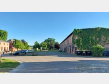 Weitläufiger Hof mit gepflastertem Boden, umgeben von niedrigen, historischen Gebäuden; rechts ein langes Backsteingebäude mit dichter Begrünung und links beige und rote Gebäude mit großen Fenstern. Im Hintergrund stehen Bäume und geparkte Autos.