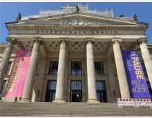 Das Bild zeigt die Fassade des Konzerthauses Berlin mit klassizistischer Architektur, bestehend aus einer Säulenreihe und einem Giebel, der mit Figuren verziert ist. Vor dem Gebäude befinden sich breite Treppen, und links sowie rechts hängen große Banner in Pink und Lila mit Hinweisen auf Veranstaltungen.