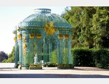 Metallpavillon im Rokoko-Stil mit grünem Gitterwerk und goldenen Verzierungen, einschließlich einer zentralen goldenen Sonnen-Dekoration; umgeben von Bäumen und Sträuchern in einer Parkanlage.