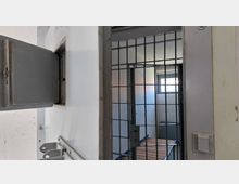 Eine Gefängniszelle mit einer grauen Eisentür, einem Ausgabeschlitz und einem vergitterten Durchgang, dahinter ein kleiner Raum mit weiß verputzten Wänden und einem vergitterten Fenster.