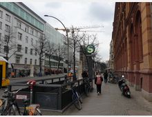 Straßenszene mit einem S-Bahn-Eingang, gekennzeichnet durch ein grünes "S"-Schild, umgeben von geparkten Fahrrädern. Rechts ein rotes Backsteingebäude mit Rundbogenfenstern, links moderne weiße und glasverkleidete Gebäude sowie eine gelbe Straßenbahn. Im Hintergrund sind Baukräne und kahle Bäume zu sehen.
