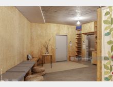 Ein minimalistischer Innenraum mit Wänden aus hellem Sperrholz und einer Betondecke. Zu den Elementen gehören eine Sitzbank mit grauen Kissen, runde geflochtene Poufs, ein kleiner Tisch mit einer Vase, ein graues Bodentür mit Regalen daneben und ein Vorhang mit Blattmuster am rechten Bildrand.