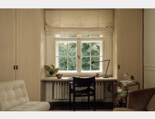 Arbeitsbereich in einem hellen Raum mit einem großen Fenster, das auf grüne Bäume blickt. Ein schlichter Holzschreibtisch mit einer schwarzen Lampe, einem Stuhl und dekorativen Elementen steht vor einer Heizung, umgeben von eingebauten Holzschränken.