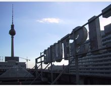 Ansicht von einem Dach in einer Stadt, mit einem vertikalen Funk- oder Fernsehturm (Fernsehturm) im Hintergrund. Im Vordergrund befindet sich die Rückseite eines stark verwitterten Schriftzuges auf einer Stahlkonstruktion, daneben sind Gebäude und ein Kran sichtbar.