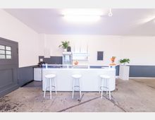 Ein heller, minimalistischer Raum mit einer weißen Theke, drei weißen Barhockern und grauem Betonboden. Im Hintergrund stehen ein Kühlschrank, Regale mit Dekoration und Pflanzen sowie eine Schale mit Orangen.