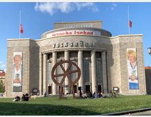 Die Aufnahme zeigt die Fassade der Volksbühne Berlin, einem monumentalen Sandsteingebäude mit hohen Säulen und dem Schriftzug "Volksbühne" über dem Eingang. Im Vordergrund steht eine große Metallskulptur, umgeben von Menschen, die auf einer Wiese sitzen; links und rechts der Fassade befinden sich vertikale Banner mit Porträts von Personen.