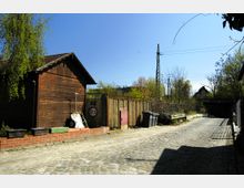 Ein gepflasterter Hofbereich mit einem Holzhäuschen links, einer Ziegelsteinmauer sowie Metallzaun im Hintergrund. Auf der rechten Seite stehen Mülltonnen und eine zweiachsige Schubkarre, umgeben von sporadischem Grün und Bäumen. Im Hintergrund sind Bahngleise und eine Eisenbahnbrücke zu sehen.