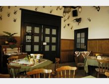 Ein rustikaler Gastraum mit Holztischen und -stühlen, an den Wänden dekoriert mit Hirschgeweihen und einem ausgestopften Vogel. Im Hintergrund sind dunkle Holztüren mit Glaseinsätzen sowie eine Holzbank mit dekorativen Kissen zu sehen.