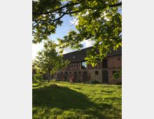 Langer Backstein-Bauernhof mit dunklem Satteldach und mehreren Türen und Fenstern, teils mit Holzdetails. Davor liegt eine grasbewachsene Wiese mit Bäumen, deren Blätterdach den Blick rahmt und Schatten auf den Rasen wirft.